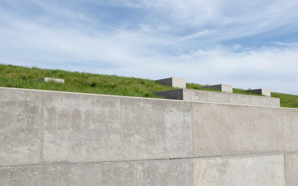 A sturdy concrete retaining wall stands prominently in the foreground, showcasing its textured surface with visible aggregate and reinforcement details. We can see the wall at a slight angle, emphasizing its height and structural integrity. In the middle ground, a gently sloping landscape features lush green grass and a few strategically placed concrete blocks to illustrate variations in design. The background presents a serene, blue sky with soft, wispy clouds, creating a calm and stable atmosphere. The lighting is bright but diffused, casting soft shadows that enhance the wall’s contours. The overall mood is one of strength and reliability, reflecting the critical qualities of concrete blocks for retaining walls. No people are present in the scene, ensuring a focus solely on the architectural features.